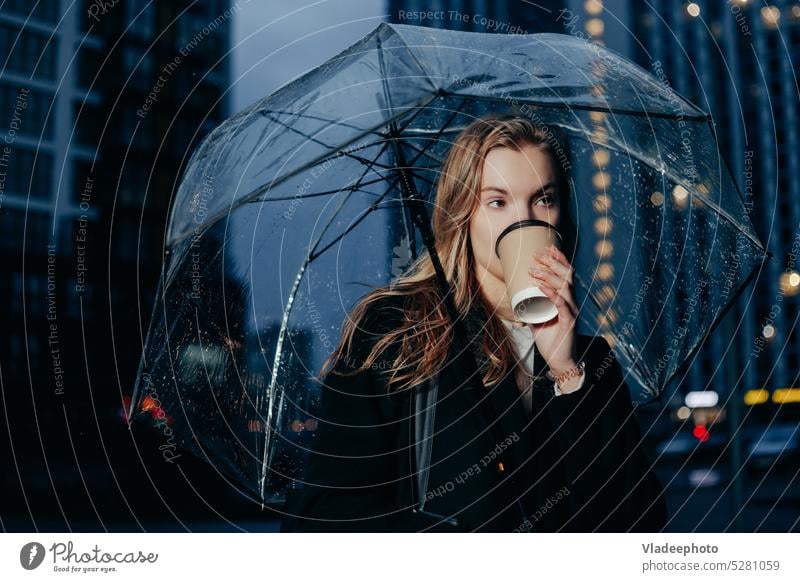 Blonde Woman in Coat Walking Under Umbrella and Holding Paper Cup in City umbrella woman cup rain holding standing paper weather autumnal night cold coffee coat