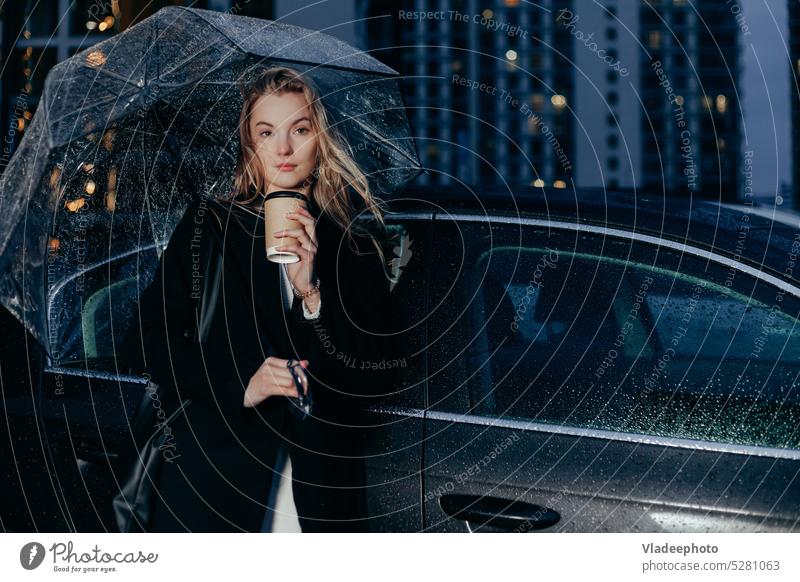 Woman stands near her car on a rainy evening under an umbrella. Drinking take away coffee woman young lady night wait taxi rent elegant access problem cheerful