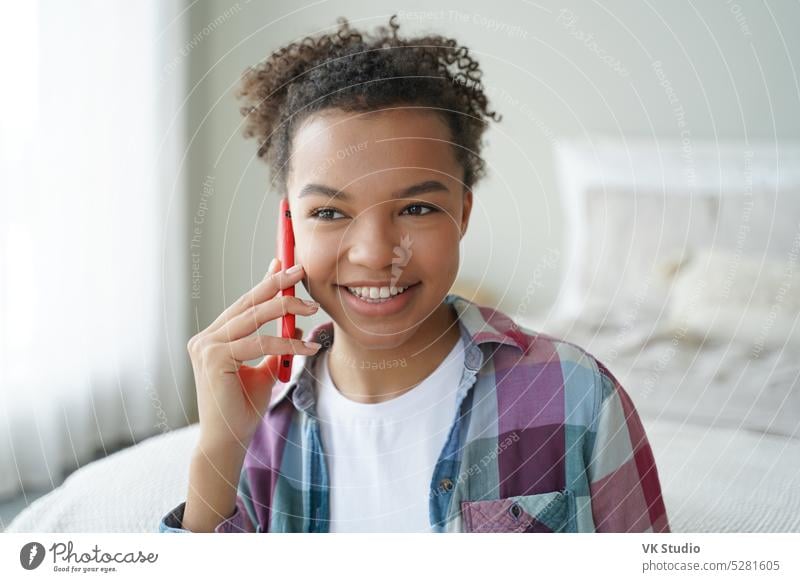Smiling mixed race teen girl answers phone call, enjoying talk with friend in bedroom at home smartphone communicate biracial speak chat outgoing optimistic