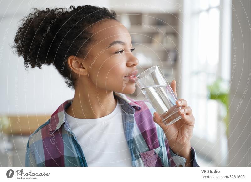 Mixed race young girl drink pure water from glass at home. Healthy lifestyle, morning routine biracial wellness hold afro diet slimming thirst teen sip purified