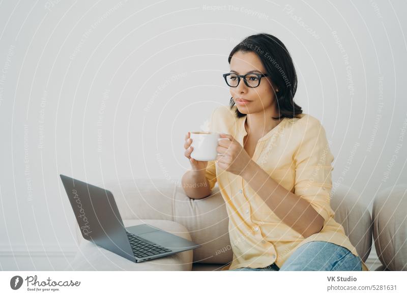 Pensive female businessperson work online on laptop holding mug of coffee at home office. Remote job e commerce drink think cup break ponder dream rest