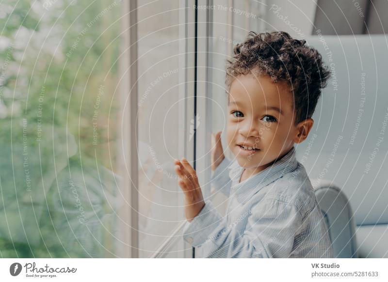 Cute mixed race little curly haired boy in light blue colored shirt next to large window kid looking son home wait parent toddler touching afro american