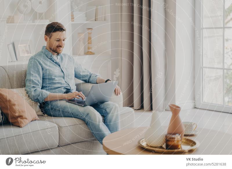 Young attractive smiling freelancer working on comfortable sofa at home man working remotely online laptop sitting cozy guy technology internet happy enjoy