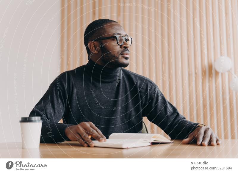 Thoughtful african businessman sits at desk with pen and agenda looks away with pensive expression young thoughtful american note book think sitting male
