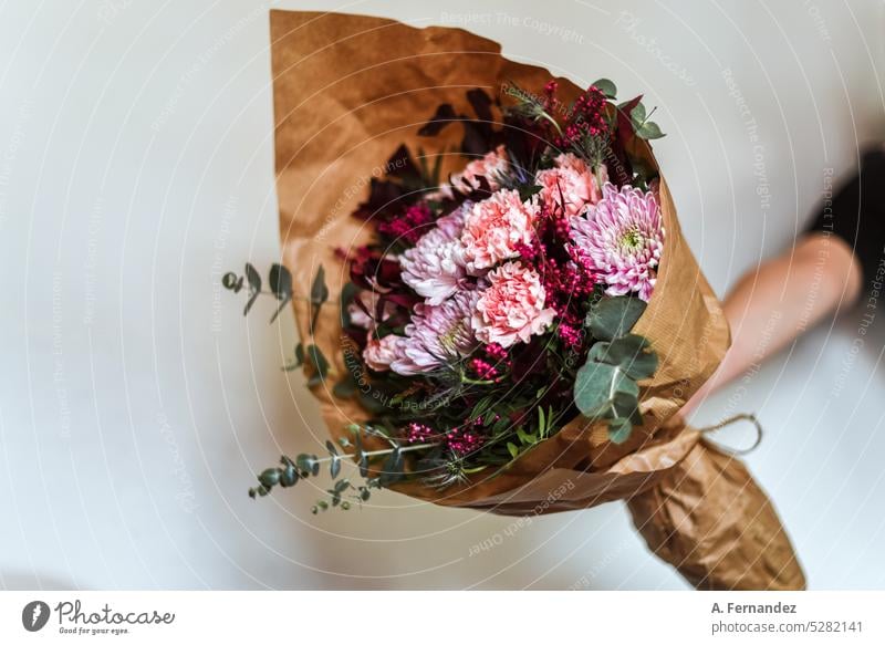 A hand with a bouquet of flowers in shades of pink and maroon, wrapped in brown paper. Silver eucalyptus, chrysanthemum, carnation, peony and blue bait. Flowers gift concept