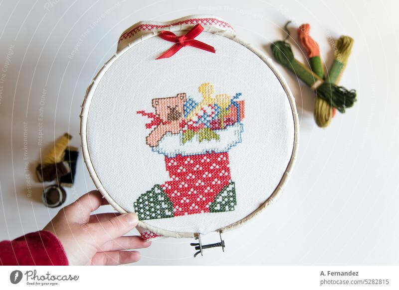 Hand holding an embroidery hoop with a piece of fabric with a cross-stitched christmas motive. Needle with a red thread. Stocking full of presents. Embroidery