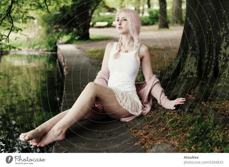 Young woman with pink blonde hair and long legs sits on the fortified shore of a lake under a tree and looks over the lake Woman portrait pretty Slim long hairs