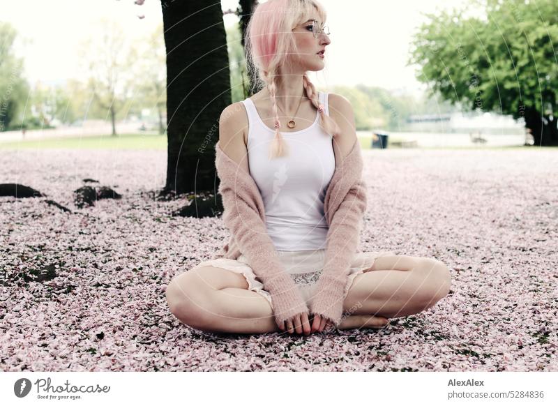Young woman with pink blonde hair sits cross-legged barefoot on the ground of a park covered with pink cherry blossom petals Woman portrait pretty Slim