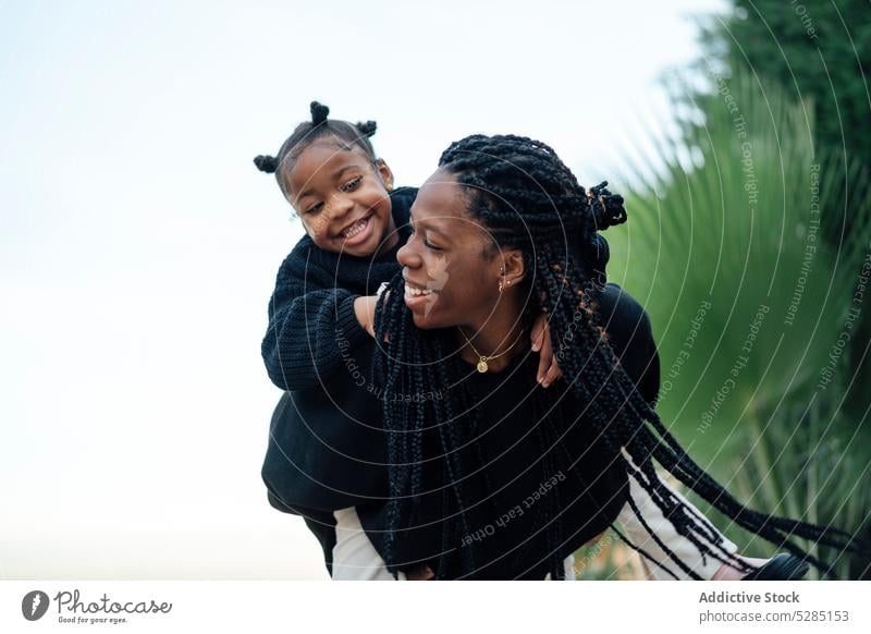 Happy black mother giving piggyback ride to daughter woman girl having fun tropical childhood weekend smile happy cheerful female kid together summer