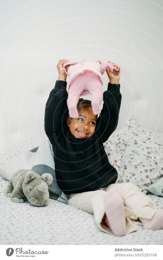 Happy little black girl sitting on bed playing with toy doll activity fun joy having fun entertain happy smile kid child african american ethnic playful
