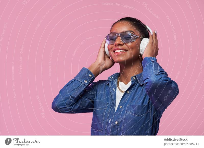 Smiling black woman in headphones listening to music on pink background trendy style fashion outfit denim sunglasses african american female young ethnic