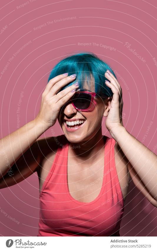 Cheerful young woman with blue hair on pink background touch head studio shot sunglasses joy enjoy carefree touch hair individuality expressive excited laugh