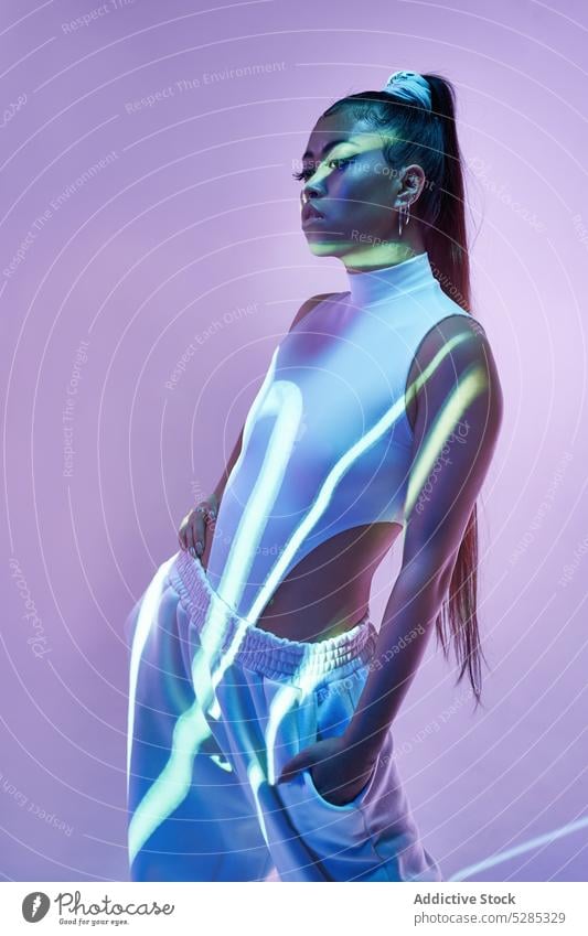 Mixed race woman in studio with neon illumination and lines from projector model hand on waist hand in pocket posture emotionless studio shot unemotional lady