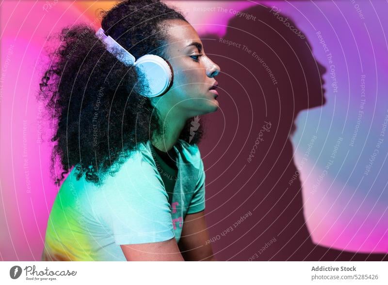 Thoughtful black woman listening to music in neon headphones meloman dreamy afro song playlist wireless light pensive hairstyle african american device gadget