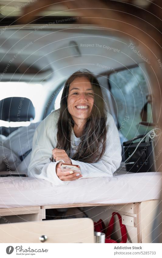 Positive young ethnic woman using smartphone and smiling in camper van lying cheerful smile road trip traveler positive sms vacation happy female hispanic