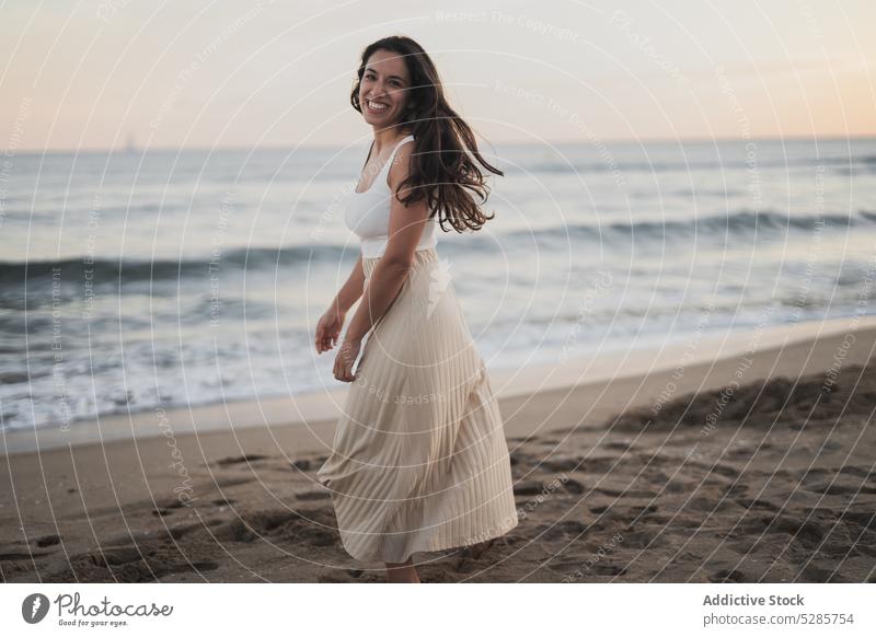 Cheerful young ethnic woman walking on sandy seashore during summer vacation beach smile enjoy happy holiday sunset traveler nature female hispanic barefoot