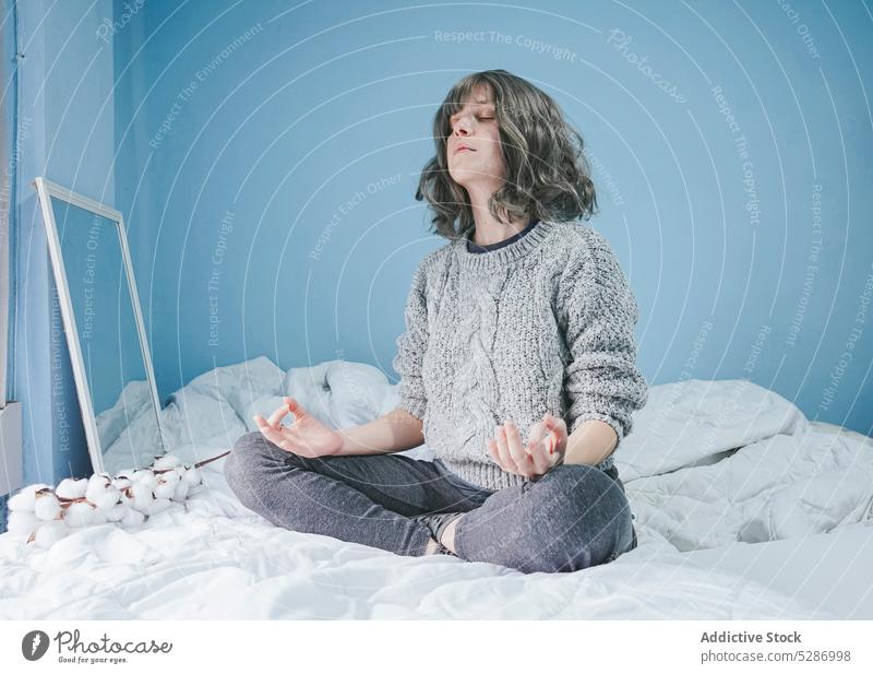 Woman in sweater meditating on bed woman meditate yoga lotus pose eyes closed practice zen bedroom warm clothes legs crossed wall calm comfort padmasana female