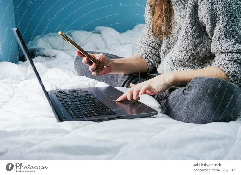 Crop woman browsing laptop and smartphone on bed using cellphone touchpad freelance self employed independent female chat project gadget online connection