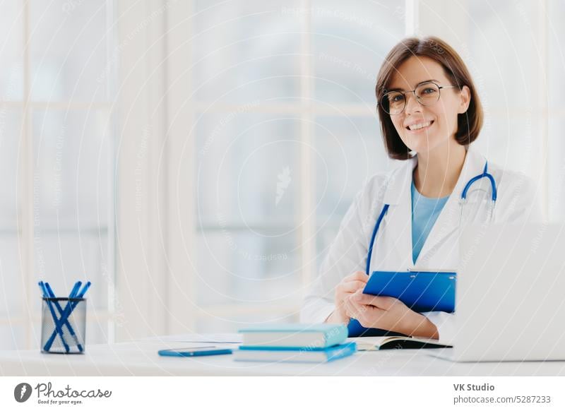 Female doctor writes prescription on special form, works in private clinic, wears white medical gown, ready for seeing patients, poses at workplace. Smiling physician or medical worker holds clipboard