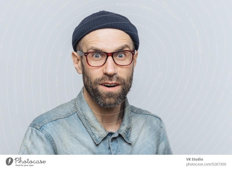 Headshot of good looking unshaven male with blue eyes, looks with suprised expression into camera, feels excited and shocked with latest news, poses against grey background. Surprisment and people