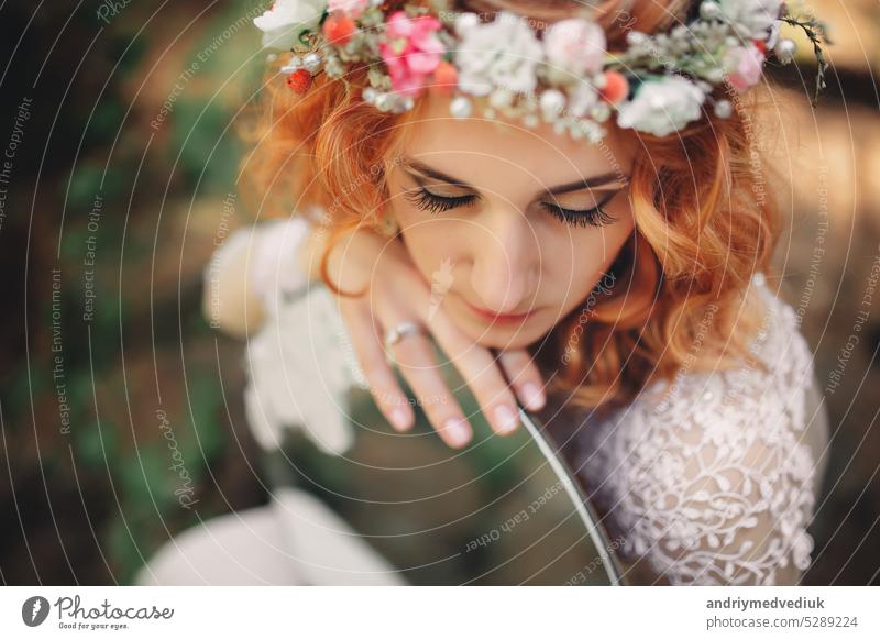 beautiful young red-haired bride in the forest with a floral wreath on her head and mirror . woman in long white dress outdoors on summer day. wedding day woods