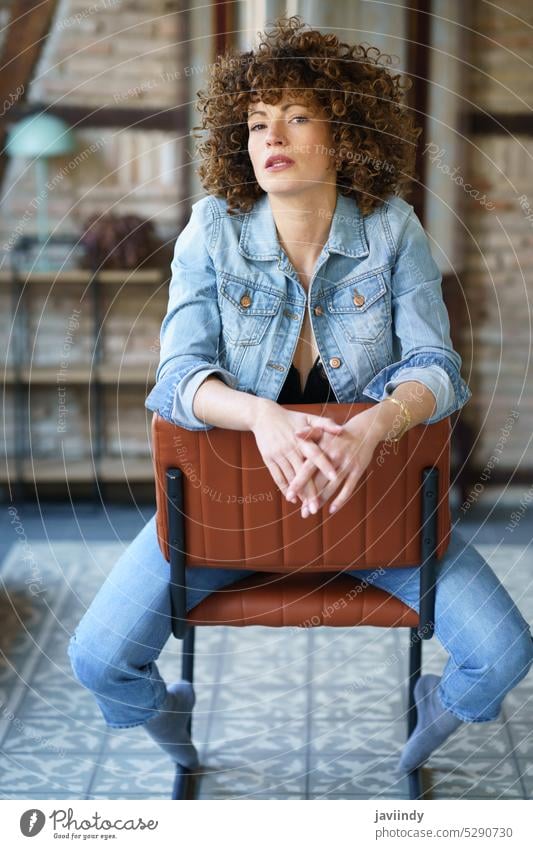 Serious young woman wearing denim jacket sitting on leather chair serious touch hair unemotional curly hair lingerie jeans appearance style room brick wall cool