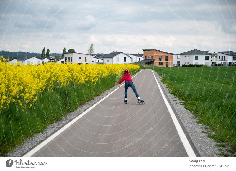 A child is inline skating on a freshly asphalted path between meadows and rape fields in the direction of a new housing estate. Sports Inline skates