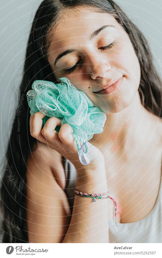 Cheerful woman holding and using an exfoliating sponge resting her face. Young adult washing her body with bath loofah. Washcloth body scrub, closeup hands rub skin in bath. Personal hygiene concept.