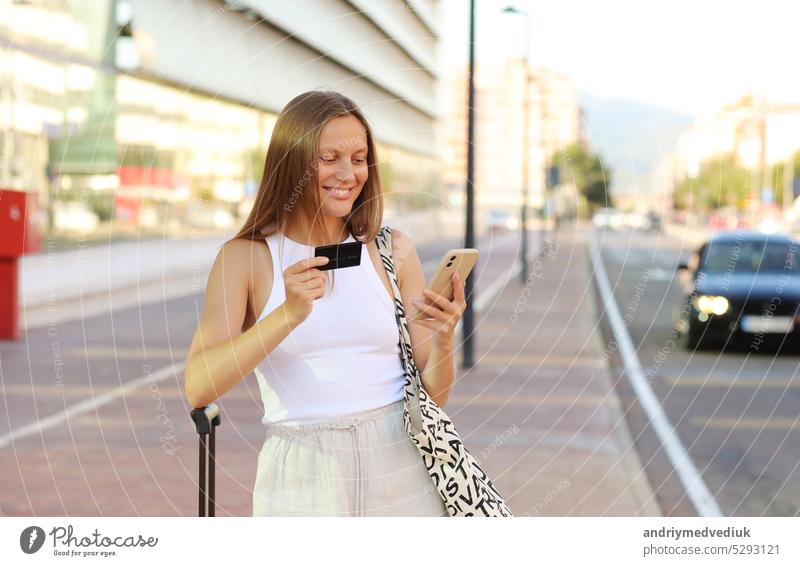 Beautiful young smiling woman with smartphone and credit card by the airport. Happy girl with suitcase after arrived airplane booking and paying for hotel. summer vacation. new abroad journey travel