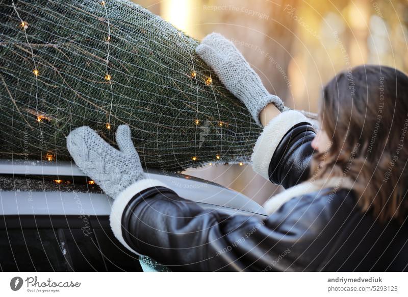 Woman packing Christmas tree with net and garlands on a rooftop of her car, getting ready for a holidays. Idea of Christmas mood and celebration. Woman wearing winter coat and knitted mittens.