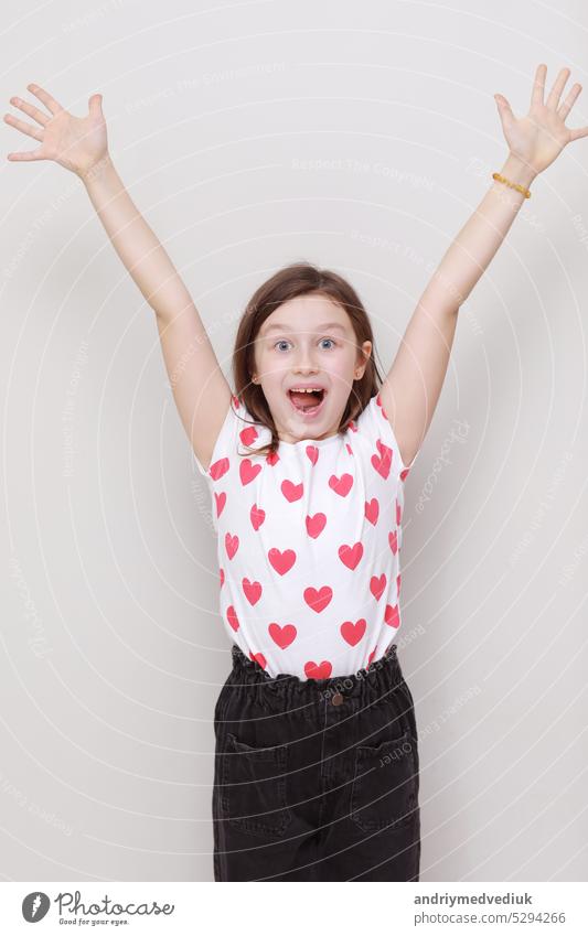 Cute smiling kid girl in white t-shirt with red hearts and stylish black jeans happily raises his hands up on white background. valentines day little isolated