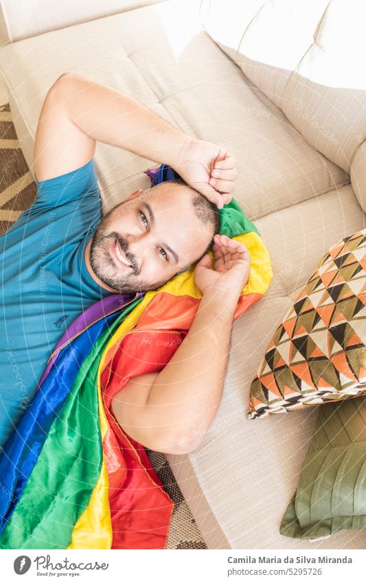 Gay man sitting on sofa at home with lgbt flag. Show less beard man bisexual equal face freedom gay gay flag gay men gay pride guy hand flag happy happy guy