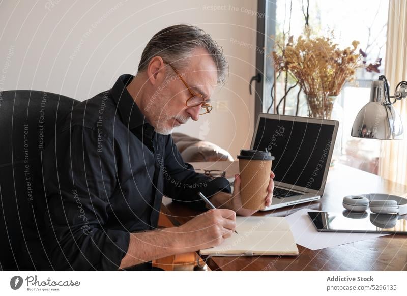 Focused freelancer writing in organizer at home office desk man write take note work notebook planner coffee concentrate table workplace hot drink male busy