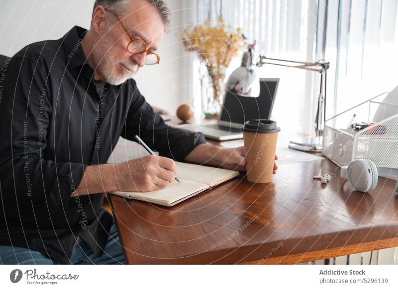 Focused freelancer writing in organizer at home office desk man write take note work notebook planner coffee concentrate table workplace hot drink male busy