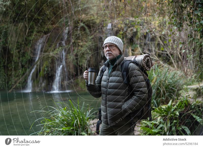 Pensive senior man with thermos and backpack forest trip hiker traveler nature enjoy mountain lakeside trekking male thoughtful warm clothes explore elderly