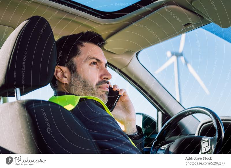 Serious male worker speaking on smartphone in car man using phone call vehicle wind turbine alternative energy cloudless sky blue sky sustainable talk wireless