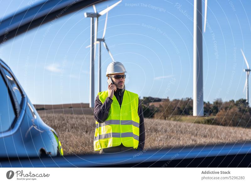 Serious male worker speaking on smartphone near car man using phone call window vest helmet vehicle turbine alternative energy cloudless sky blue sky