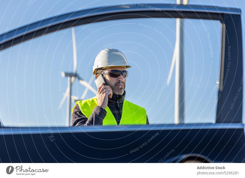 Serious male worker speaking on smartphone near car man using phone call window vest helmet vehicle turbine alternative energy cloudless sky blue sky