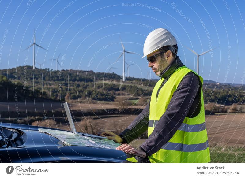 Male engineer examining map on hood of car man examine countryside worker wind turbine innovation power alternative energy cloudless sky sustainable propeller