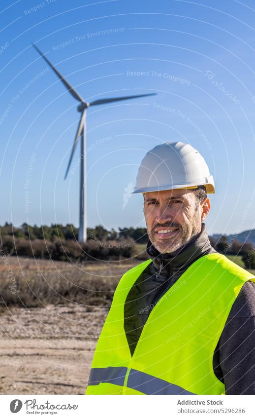 Smiling engineer standing in countryside field against windmill man turbine work alternative energy eco friendly power positive smile cloudless sky ecosystem