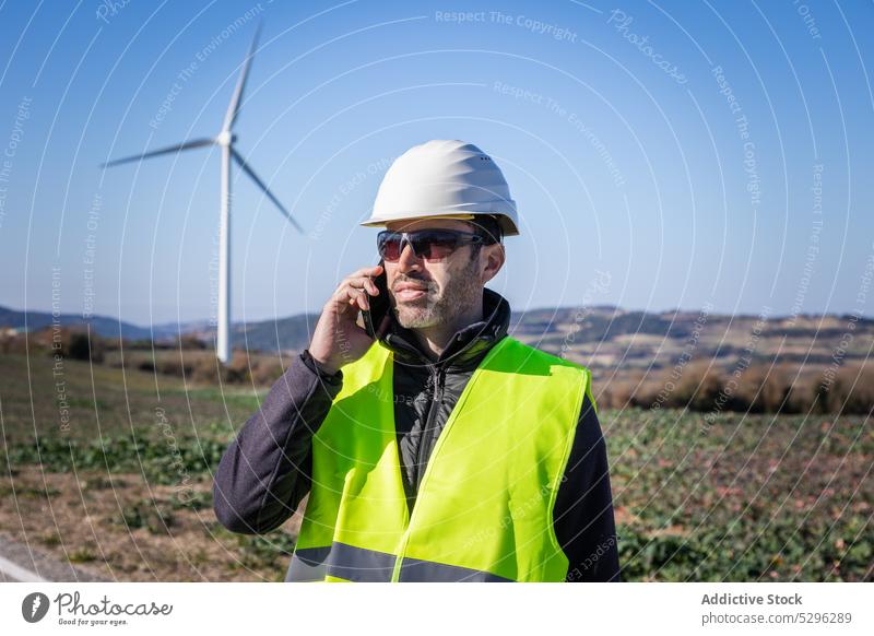 Engineer in protective helmet speaking on smartphone against wind turbines man engineer countryside roadway worker using phone call eco alternative energy