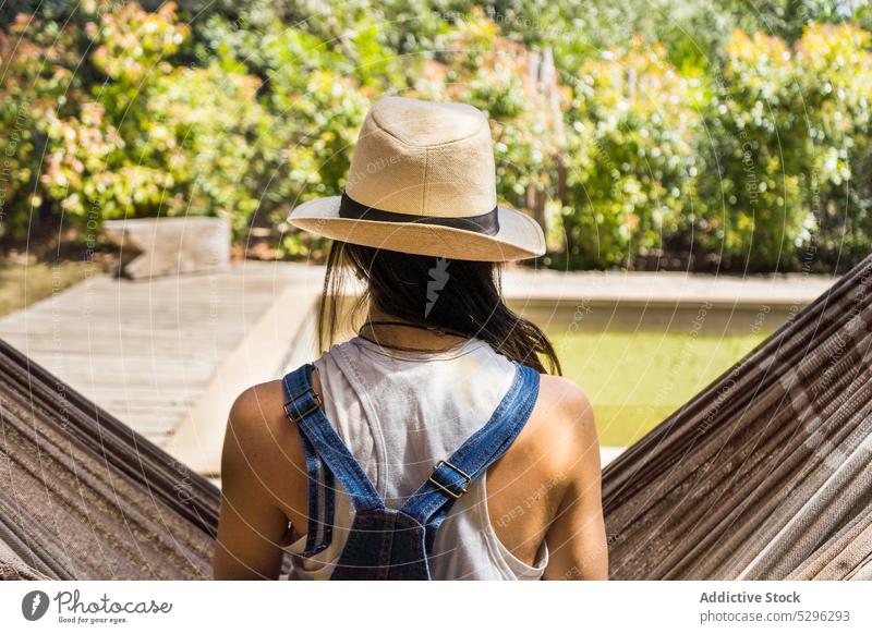 Unrecognizable woman in hat resting on hammock pool relax resort weekend summer vacation holiday tree sun daytime female recreation nature chill leisure sit