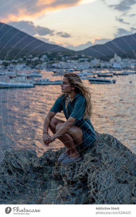 Young lady sitting on boulder on seashore during summer holidays woman tourist beach sunset rest vacation thoughtful calm coast female young blond long hair