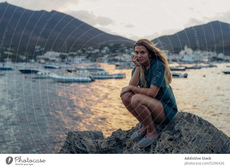Young lady sitting on boulder on seashore during summer holidays woman tourist beach sunset hand at chin rest vacation thoughtful calm coast female young blond