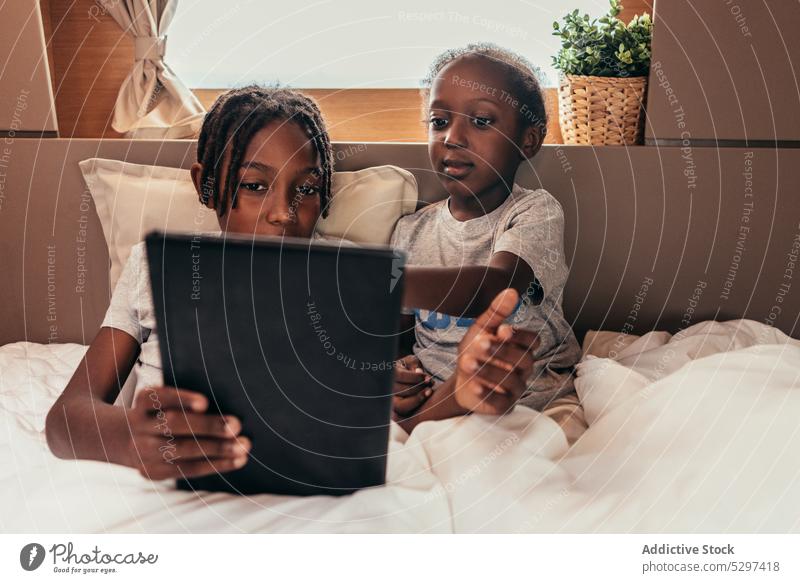 Content African American children watching movie on bed together tablet blanket rest using weekend relax gadget lying device internet comfort sibling video