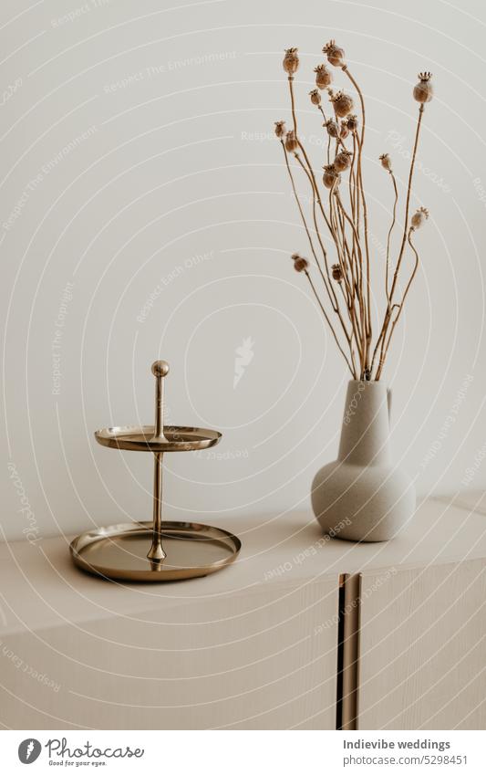 A golden plated cake stand and a beige ceramic vase with dried flowers on the cabinet. Copy space, white background. Modern home decoration. bouquet spring
