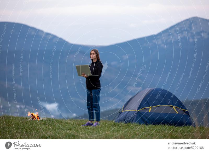 young female freelancer working on laptop in the mountains in the evening. Tourist girl sitting near campfire and having fun. Copy space. tourist online travel