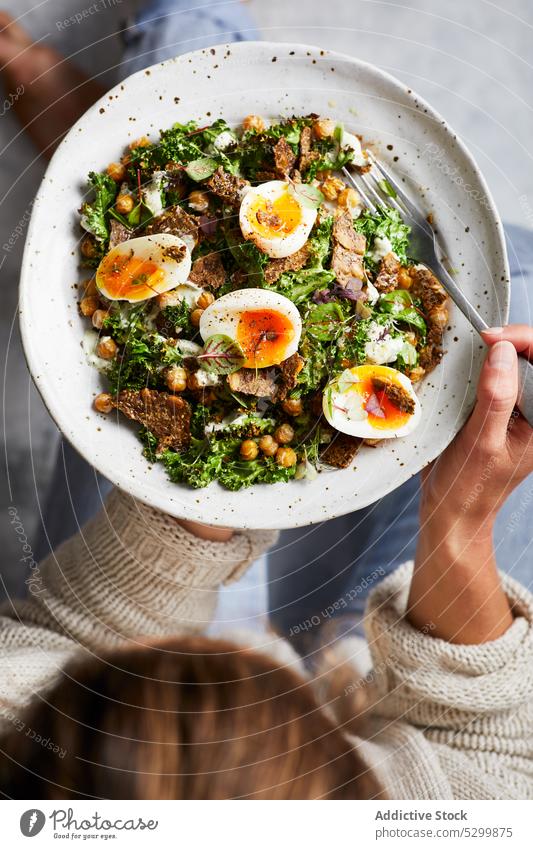 Crop anonymous woman eating appetizing salad with eggs and chickpeas dish healthy food lunch hungry herb female young sweater tasty yummy kitchen daylight