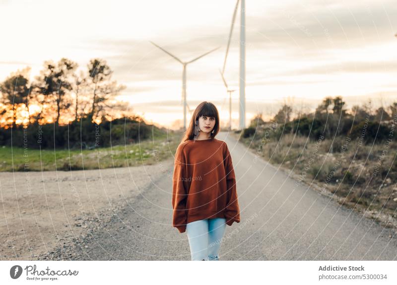 Calm woman walking along road in countryside traveler confident sunset windmill nature trip young female tree casual serious style almatret catalonia spain