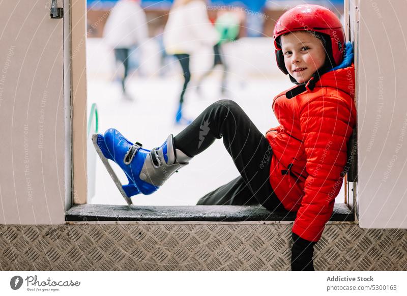 Boy in skates sitting on border of ice rink boy winter safety step kid learn leisure sport activity hobby cold snow focus recreation active weekend warm clothes